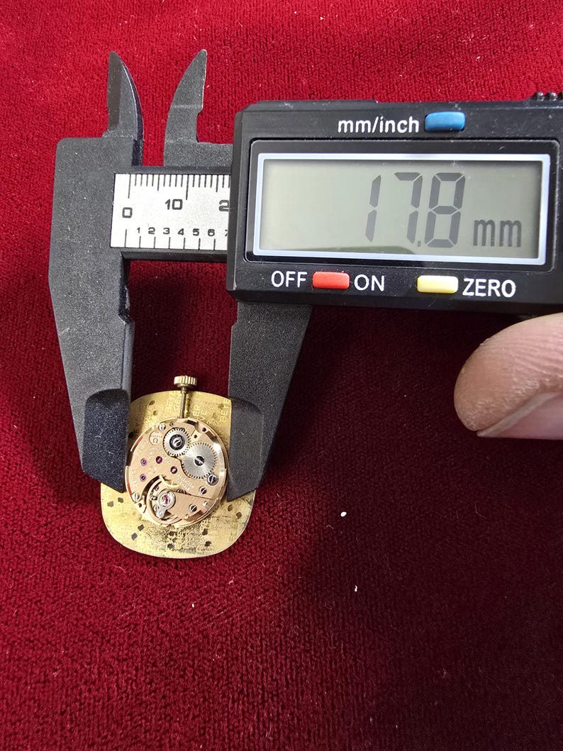 Pu&ograve; includere: Un movimento di orologio dorato, di circa 1,78 cm di diametro, viene misurato con un calibro digitale. Il meccanismo dell'orologio presenta ingranaggi intricati e rubini. Il calibro mostra la misura in millimetri.