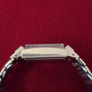 May include: A gold-toned rectangular wristwatch with a silver and gold band. The watch face is rectangular and has a clear crystal. The band is made of small, interlocking metal links. The watch is set against a red background.