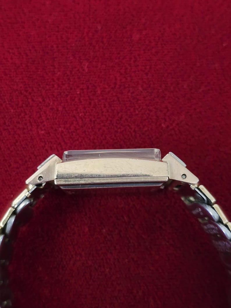 May include: A gold-toned rectangular wristwatch with a silver and gold band. The watch face is rectangular and has a clear crystal. The band is made of small, interlocking metal links. The watch is set against a red background.