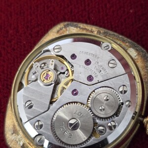 May include: Close-up of a vintage gold-toned watch with the back removed, revealing the intricate mechanical movement. The watch mechanism features silver-colored gears, jewels, and the text "SEVENTEEN 17 JEWELS SWISS UNADJUSTED".