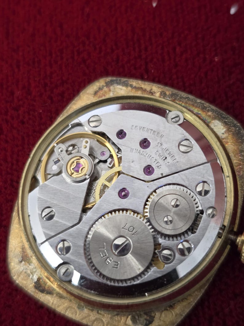 May include: Close-up of a vintage gold-toned watch with the back removed, revealing the intricate mechanical movement. The watch mechanism features silver-colored gears, jewels, and the text "SEVENTEEN 17 JEWELS SWISS UNADJUSTED".