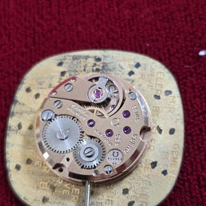 May include: Close-up of a vintage Omega watch movement. The gold-toned mechanism features intricate gears, jewels, and the Omega logo. The watch movement is mounted on a gold-colored base with text around the edges. The background is a deep red fabric.