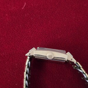 May include: A gold-toned rectangular wristwatch with a silver and gold band. The watch face is rectangular with a small round dial. The watch band is a mix of silver and gold links. The watch is set against a red background.