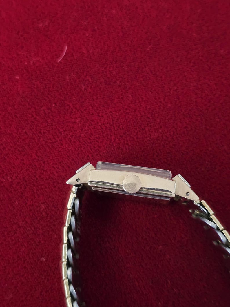 May include: A gold-toned rectangular wristwatch with a silver and gold band. The watch face is rectangular with a small round dial. The watch band is a mix of silver and gold links. The watch is set against a red background.