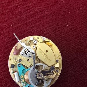 Puede incluir: Primer plano de un mecanismo de reloj con engranajes, tornillos y muelles intrincados. El mecanismo dorado presenta componentes plateados, turquesa y cobrizos. Se puede leer el texto "Sears Roebuck & Co.".