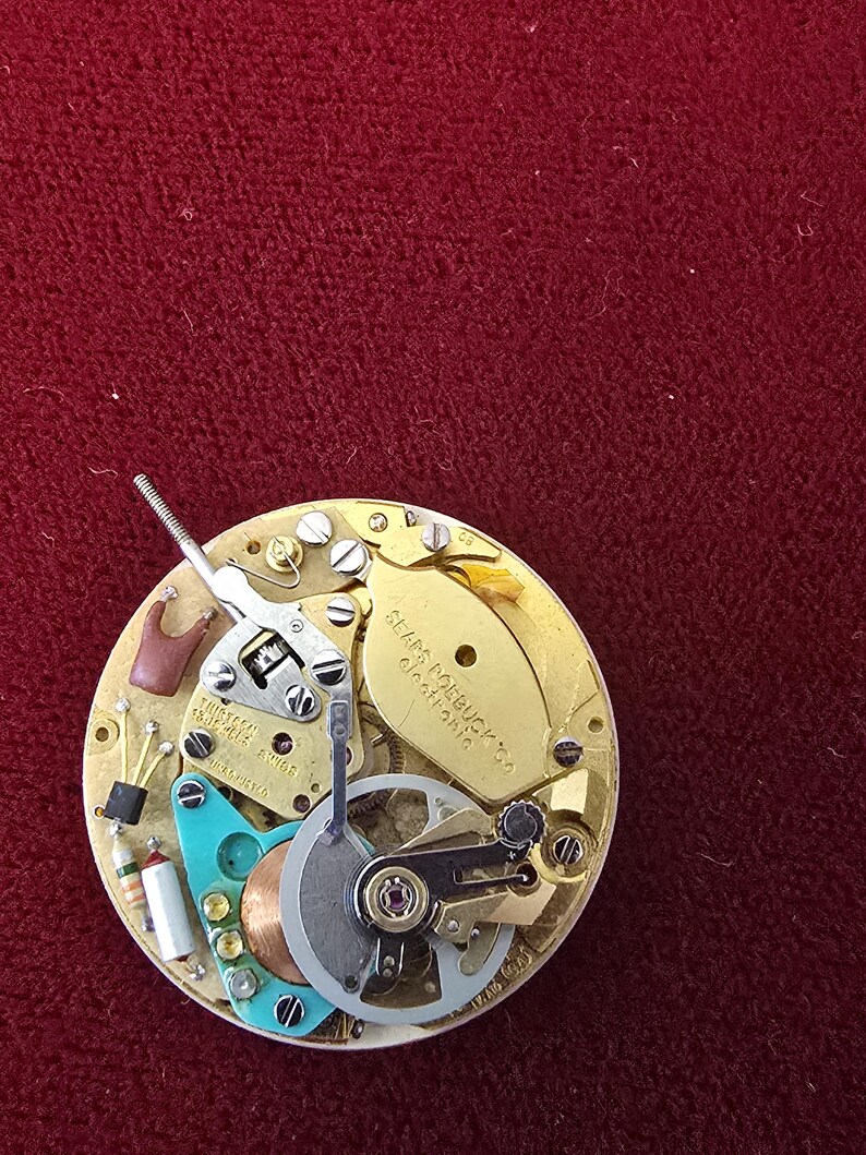 Puede incluir: Primer plano de un mecanismo de reloj con engranajes, tornillos y muelles intrincados. El mecanismo dorado presenta componentes plateados, turquesa y cobrizos. Se puede leer el texto "Sears Roebuck & Co.".