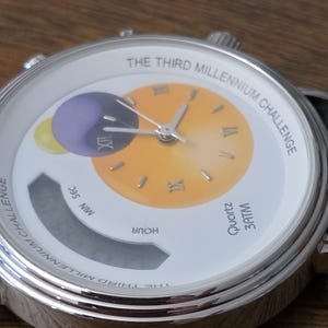 Op de afbeelding: Een zilverkleurig analoog en digitaal polshorloge met een witte wijzerplaat met de tekst THE THIRD MILLENNIUM CHALLENGE. De wijzerplaat bevat een gele gradi&euml;ntcirkel met Romeinse cijfers, een paarse en gele afbeelding en een digitaal displayvenster.