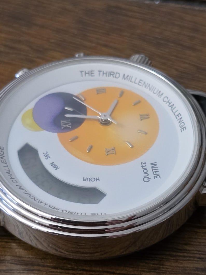 Op de afbeelding: Een zilverkleurig analoog en digitaal polshorloge met een witte wijzerplaat met de tekst THE THIRD MILLENNIUM CHALLENGE. De wijzerplaat bevat een gele gradi&euml;ntcirkel met Romeinse cijfers, een paarse en gele afbeelding en een digitaal displayvenster.
