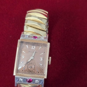 May include: A vintage Bulova watch with a gold-toned rectangular face and a gold band. The watch face features the number 12, 3, and 9, with a small seconds dial. The band has a unique design with ruby and diamond accents.