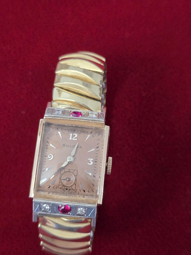 May include: A vintage Bulova watch with a gold-toned rectangular face and a gold band. The watch face features the number 12, 3, and 9, with a small seconds dial. The band has a unique design with ruby and diamond accents.