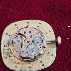 Pu&ograve; includere: Primo piano di un movimento di orologio con ingranaggi, gioielli e una corona di carica. Il movimento dorato &egrave; montato su una base dorata. Lo sfondo &egrave; rosso. Testi e marcature sono visibili.