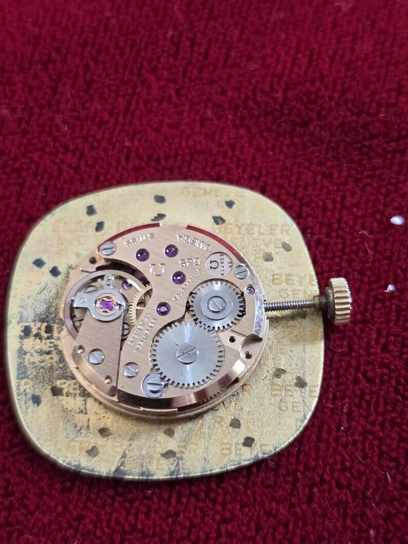 Pu&ograve; includere: Primo piano di un movimento di orologio con ingranaggi, gioielli e una corona di carica. Il movimento dorato &egrave; montato su una base dorata. Lo sfondo &egrave; rosso. Testi e marcature sono visibili.