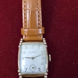 May include: Vintage Elgin wristwatch with a rectangular silver-tone case and a light-colored face. The watch features a brown leather band with a textured pattern. The watch face displays the brand name and the time.