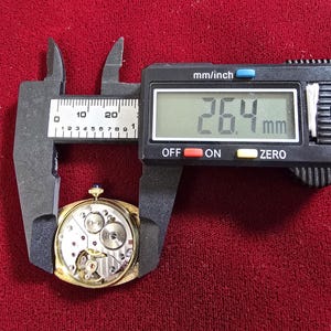 May include: A gold-toned watch movement is measured with a digital caliper. The measurement reads 26.4 mm, approximately 1.04 inches. The watch mechanism is visible, showcasing gears and intricate components. The background is a deep red fabric.