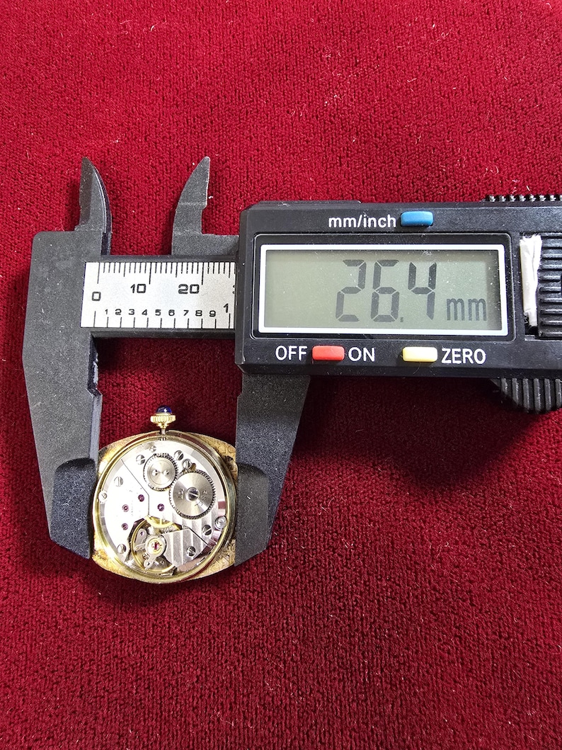 May include: A gold-toned watch movement is measured with a digital caliper. The measurement reads 26.4 mm, approximately 1.04 inches. The watch mechanism is visible, showcasing gears and intricate components. The background is a deep red fabric.