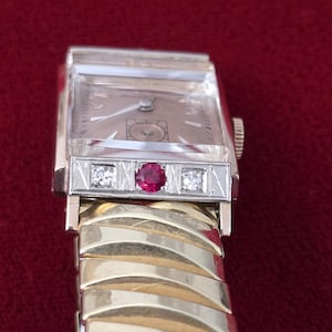 May include: Vintage gold and silver wristwatch with a rectangular face. The watch features a gold band and a silver bezel with a ruby and two diamonds. The watch face is a light tan color.