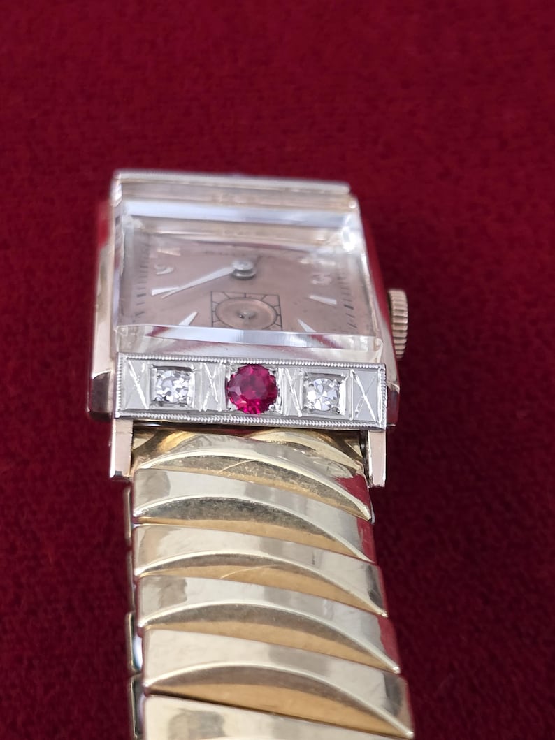 May include: Vintage gold and silver wristwatch with a rectangular face. The watch features a gold band and a silver bezel with a ruby and two diamonds. The watch face is a light tan color.