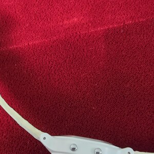 May include: A white digital watch with a cream-colored band. The watch face has two silver buttons and a clear display. The watch is set against a red textured background.