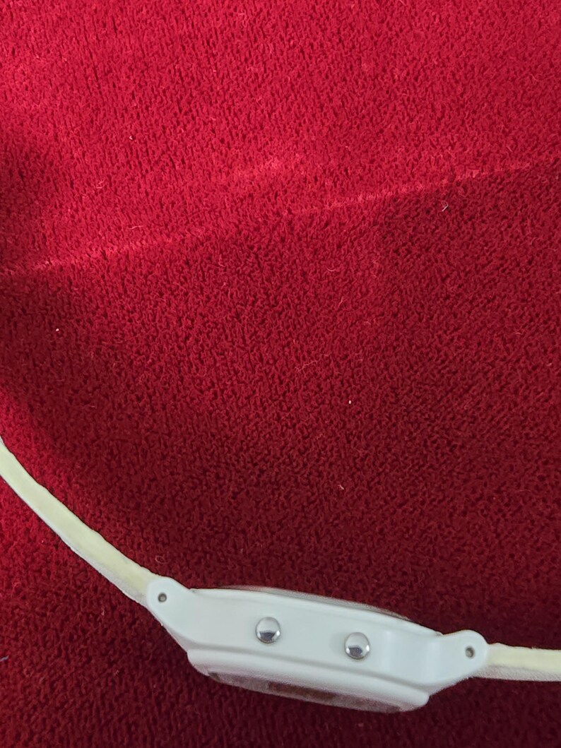 May include: A white digital watch with a cream-colored band. The watch face has two silver buttons and a clear display. The watch is set against a red textured background.