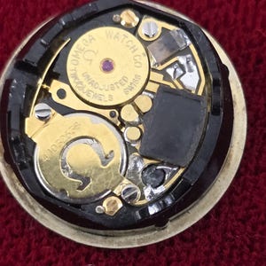 K&ouml;nnte beinhalten: Nahaufnahme eines Omega-Uhrwerks. Der Uhrenmechanismus zeigt goldfarbene Zahnr&auml;der, Schrauben und eine schwarze Komponente. Der Text "OMEGA WATCH CO SWISS" ist auf einem goldfarbenen kreisf&ouml;rmigen Teil zu sehen. Der Hintergrund ist tiefrot.