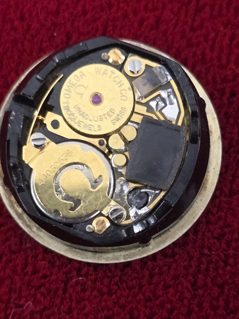 K&ouml;nnte beinhalten: Nahaufnahme eines Omega-Uhrwerks. Der Uhrenmechanismus zeigt goldfarbene Zahnr&auml;der, Schrauben und eine schwarze Komponente. Der Text "OMEGA WATCH CO SWISS" ist auf einem goldfarbenen kreisf&ouml;rmigen Teil zu sehen. Der Hintergrund ist tiefrot.