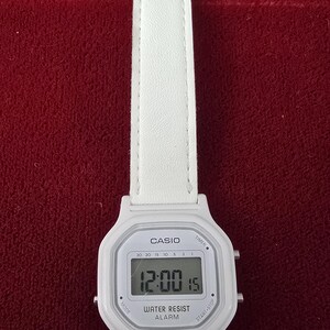 May include: White Casio digital watch with a white leather strap. The watch face displays the time as 12:00:15. Features include a timer, alarm, and water resistance.