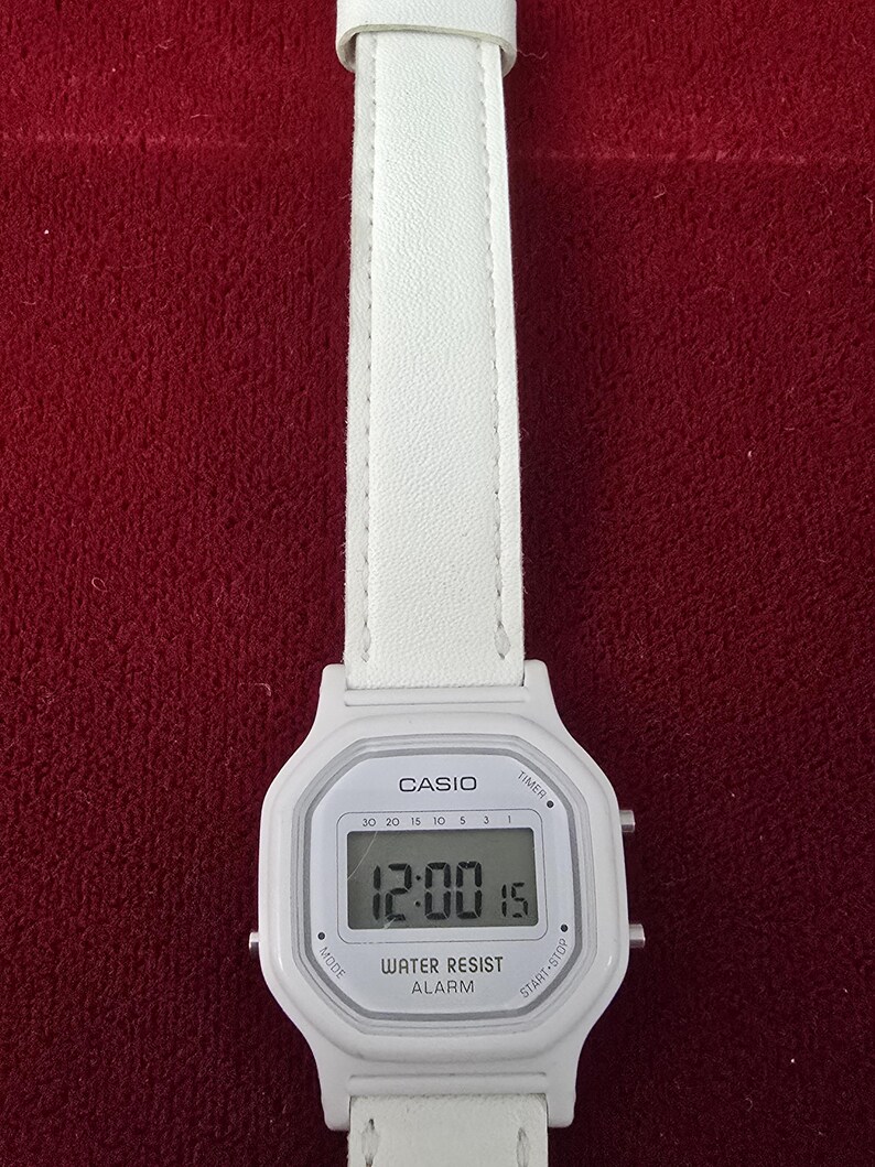 May include: White Casio digital watch with a white leather strap. The watch face displays the time as 12:00:15. Features include a timer, alarm, and water resistance.