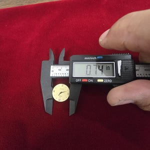 K&ouml;nnte beinhalten: Ein goldfarbenes Zifferblatt mit einem Durchmesser von etwa 1,88 cm wird mit einem digitalen Messschieber gemessen. Das Zifferblatt hat schwarze r&ouml;mische Ziffern und Zeiger. Der Hintergrund ist ein tiefroter Stoff.