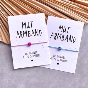 Könnte beinhalten: Zwei Karten mit dem Text "MUT ARMBAND" und "DU KANNST ALLES SCHAFFEN!". Jede Karte zeigt ein Armband mit einer Smiley-Perle. Ein Armband ist rosa, das andere blau. Der Hintergrund umfasst eine goldfarbene, fächerförmige Dekoration.