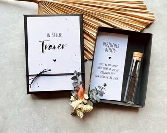 May include: Two black boxes with white cards and text. One box reads "IN STILLER Trauer" and the other reads "HERZLICHES BEILEID". A small glass vial with a cork stopper is inside one box. Dried flowers and a decorative leaf are also present.