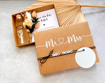May include: A brown box with a white inscription that reads "Mr & Mrs" with a heart symbol. The box contains a small glass bottle with a cork stopper and a dried flower bouquet. The box also has a white circle with a heart symbol and a black string with white beads that spell "TIM LOLA".