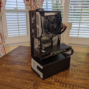 May include: A custom open-frame computer build featuring a white and silver motherboard labelled Titanium, a black CPU cooling fan with AMD branding, and a black graphics card with green GeForce GTX text, all mounted on a solid black rectangular base.