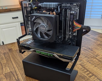May include: An open-frame computer chassis featuring a black metal stand, an ASRock B450M Pro4 motherboard, an AMD CPU cooler, two RAM sticks, and a Gigabyte GeForce GTX graphics card with visible green lettering and multi-colored power cables.
