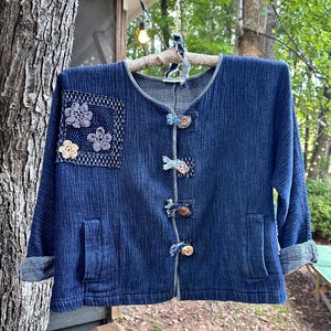 May include: A dark blue denim jacket with long sleeves and a round neckline. The front features four toggle closures with buttons, two side pockets, and a square patch on the upper left side decorated with three small, crocheted flower appliques.