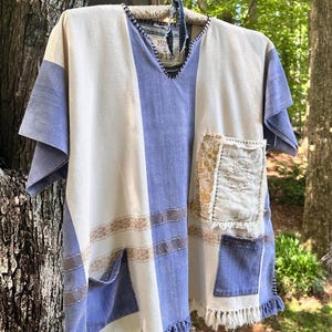 May include: A short-sleeved poncho featuring vertical panels of cream and periwinkle blue fabric with horizontal tan woven stripes. It includes a V-neckline, a fringed hem, and three patch pockets with visible stitching, including one with a floral pattern.
