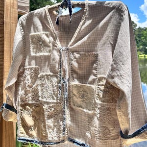 May include: A beige long-sleeve shirt with a subtle white grid pattern, featuring a V-neckline with tie closures. The front is adorned with various square patches of lace, textured fabric, and solid beige material, finished with frayed dark blue edges.