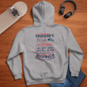 Puede incluir: Sudadera con capucha gris con un estampado gráfico que dice "Trucker's Wife" con una imagen de camión. El texto debajo dice "Sí, está trabajando. No, no sé cuándo volverá a casa. Sí, todavía estamos casados. No, no es imaginario."