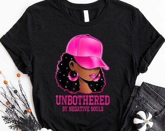 May include: Black t-shirt with a pink graphic of a woman wearing a pink hat and earrings. The text "Unbothered by Negative Souls" is printed in pink below the graphic.
