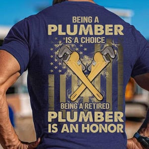 May include: Navy blue t-shirt with the text "BEING A PLUMBER IS A CHOICE, BEING A RETIRED PLUMBER IS AN HONOR." The design includes crossed wrenches and an American flag.