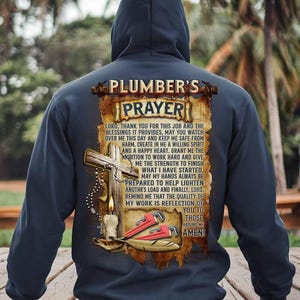 May include: Navy blue hooded sweatshirt featuring a graphic with the text "PLUMBER'S PRAYER". The graphic includes a cross, tools, and a candle. The text is a prayer for plumbers.