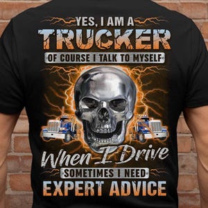 May include: Black t-shirt with a graphic featuring a chrome skull, lightning bolts, and two blue semi-trucks. Text reads "YES, I AM A TRUCKER" and "When I Drive, Sometimes I Need EXPERT ADVICE."
