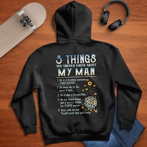 Peut inclure: Sweat-shirt à capuche noir avec un imprimé graphique blanc qui dit "5 choses à savoir sur mon homme" avec une liste de cinq choses sur un pompier. Le graphique comprend également des tournesols et un casque de pompier.