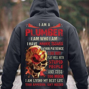 May include: A dark gray hooded sweatshirt with the text "I AM A PLUMBER" in red and white. Below the text is a graphic of a skull with red and yellow accents. The sweatshirt also has text that reads "I AM WHO I AM" and "YOUR APPROVAL ISN'T NEEDED."