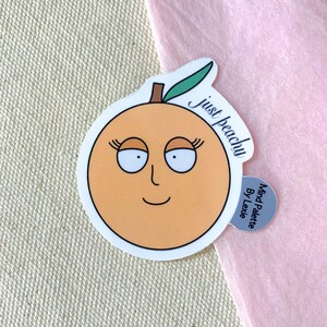 May include: A peach-colored sticker with a cartoon peach face and the text "just peachy" in black cursive. The sticker is on a light pink background. The sticker also has a small silver circle with the text "Mind Palette By Lexie" in black.