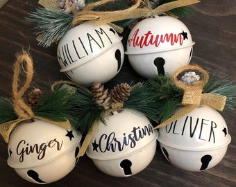 May include: Five white metal jingle bells with black and gold accents. Each bell is decorated with a different name: William, Autumn, Ginger, Christina, and Oliver. The bells are surrounded by sprigs of pine and burlap ribbon.