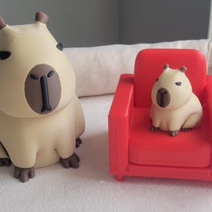 Puede incluir: Una pequeña figura de capibara beige se sienta junto a un sillón rojo con una figura de capibara más pequeña sentada en él. Ambas figuras tienen ojos y narices negros.