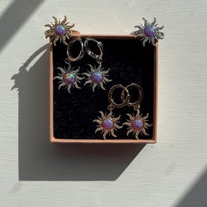 Puede incluir: Un conjunto de seis pendientes de oro y plata con un diseño de sol y piedras de color púrpura iridiscente. Los pendientes se muestran en una caja negra con tapa rosa.