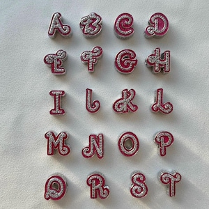May include: Set of 26 silver charms with pink enamel and clear rhinestones, each charm features a letter of the alphabet.