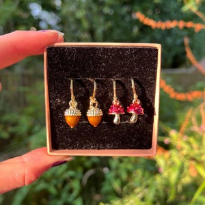 May include: A set of three gold-tone dangle earrings featuring a mushroom and acorn design. The earrings are displayed in a small, red velvet box.