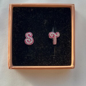 May include: A pink and silver zodiac earrings set in a box. The earrings are shaped like the zodiac signs for Sagittarius and Scorpio.